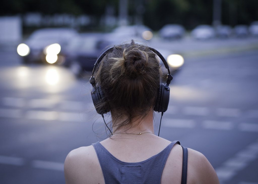 Person standing by road looking at cars, wearing headphones.
