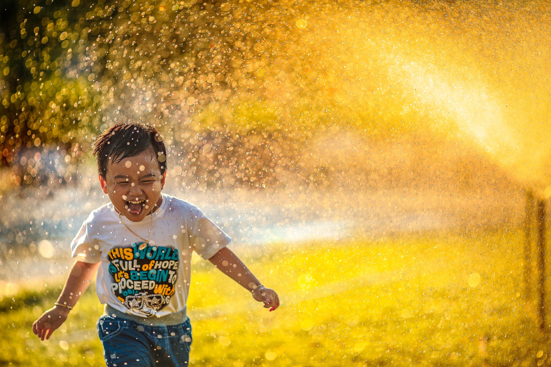 Child wearing tee shirt and jeans running through sprinkler.