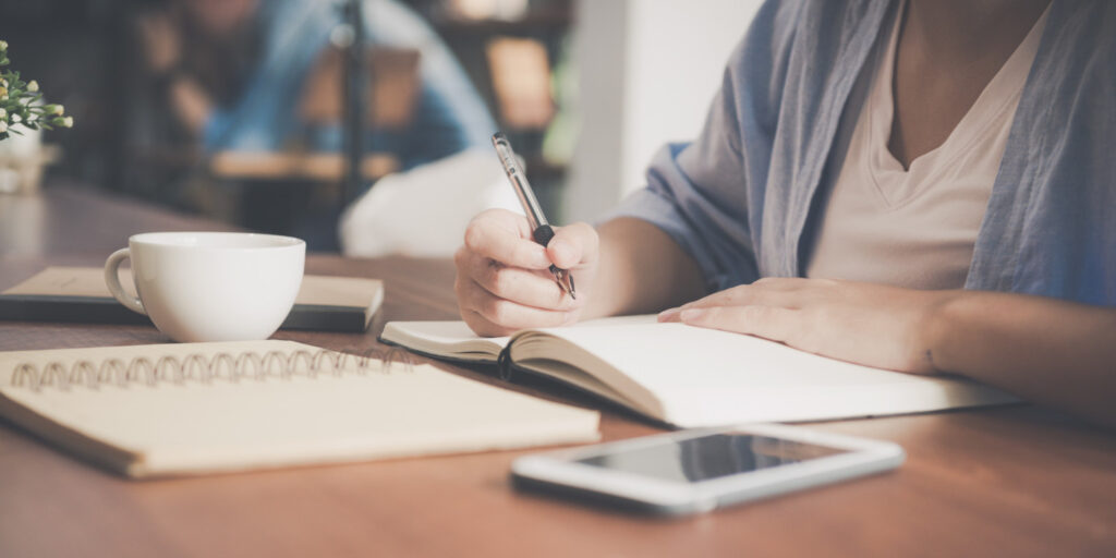 Person sitting a table with coffee, journal, phone and holding a pen writing.