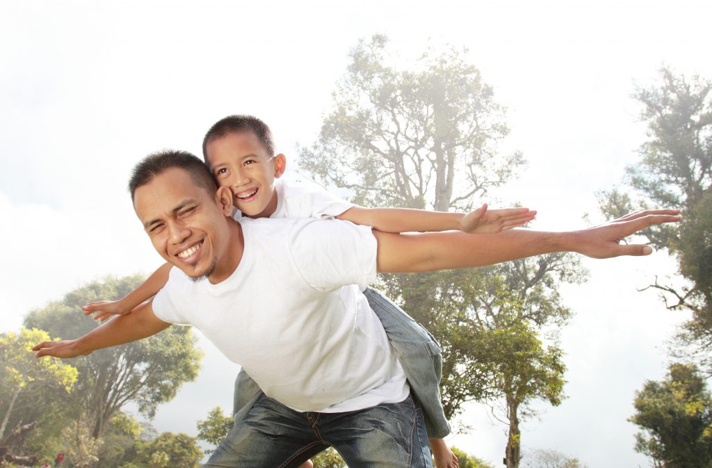 Father and son playing outside. Father is holding son up on his back and they have their arms stretched out.