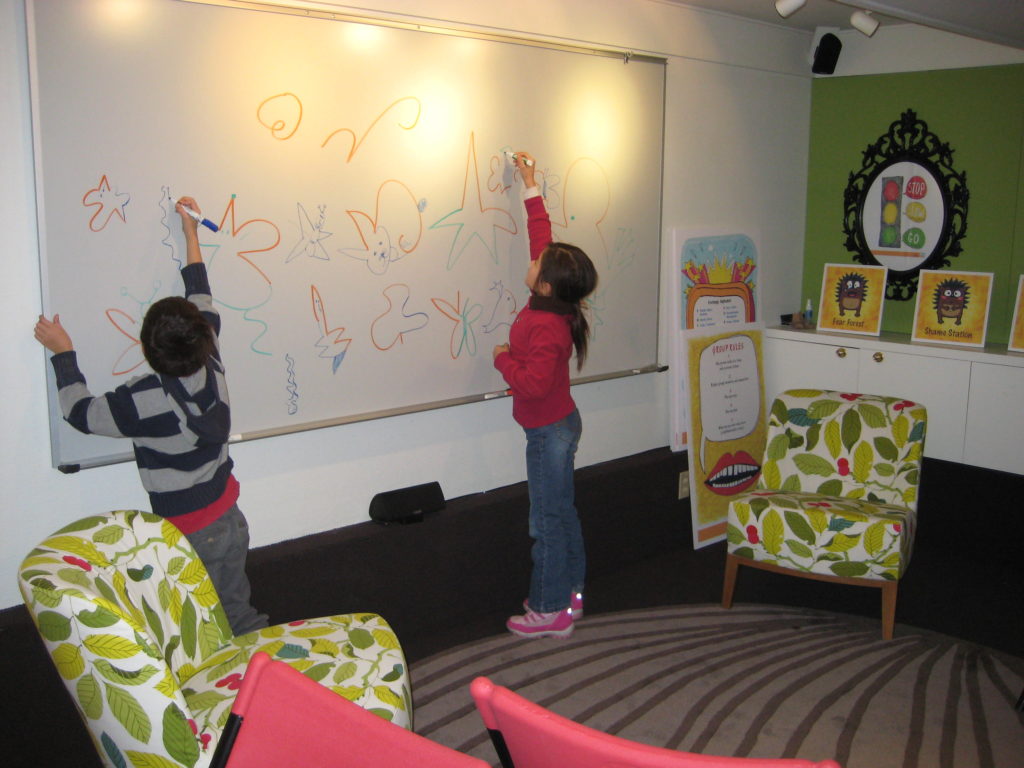 Two children drawing animals and shapes on a whiteboard.