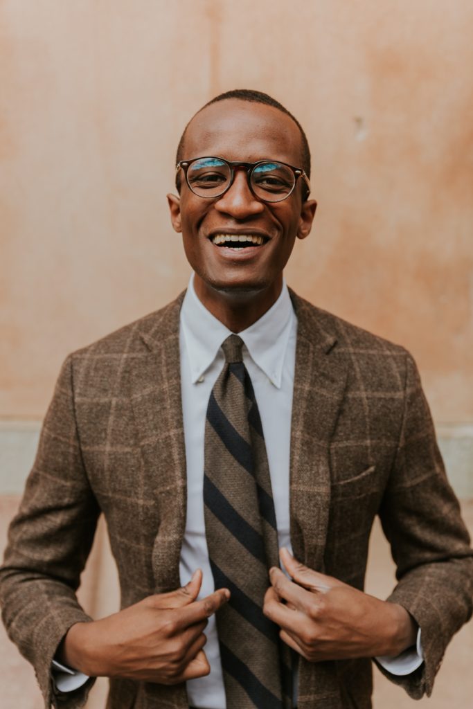A man with glasses wearing a nice brown suit and tie, smiling at the camera.