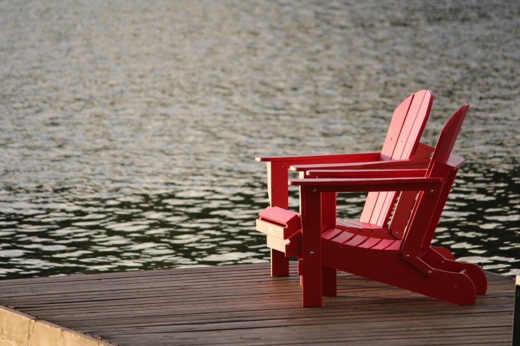 Two red Adirondack chairs on dock at a lake.